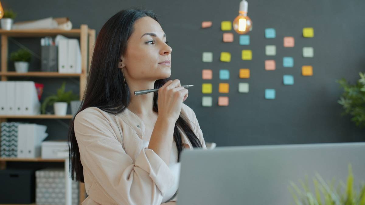 Beautiful young businesswoman is thinking then working with laptop in office, guy is busy in background. Ambitious people and occupation concept.