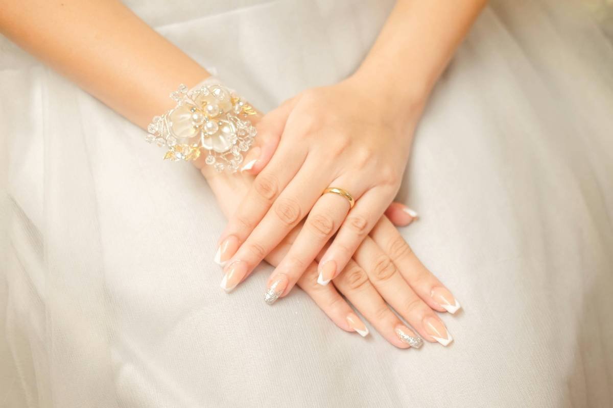 Elegant Bridal Hands with Wedding Ring. A close-up shot of a bride's hands adorned with a gold wedding ring and a delicate floral bracelet, showcasing elegant French manicure with glitter accents. Perfect for capturing the beauty and details of wedding day preparations.